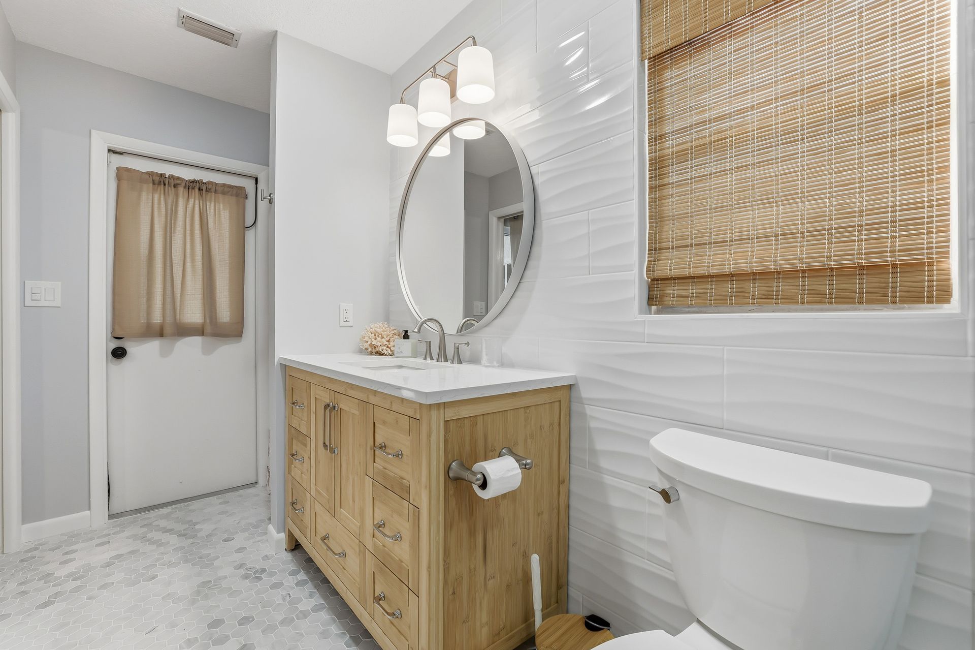 Bright bathroom with wood vanity, round mirror, white toilet, and woven window shade