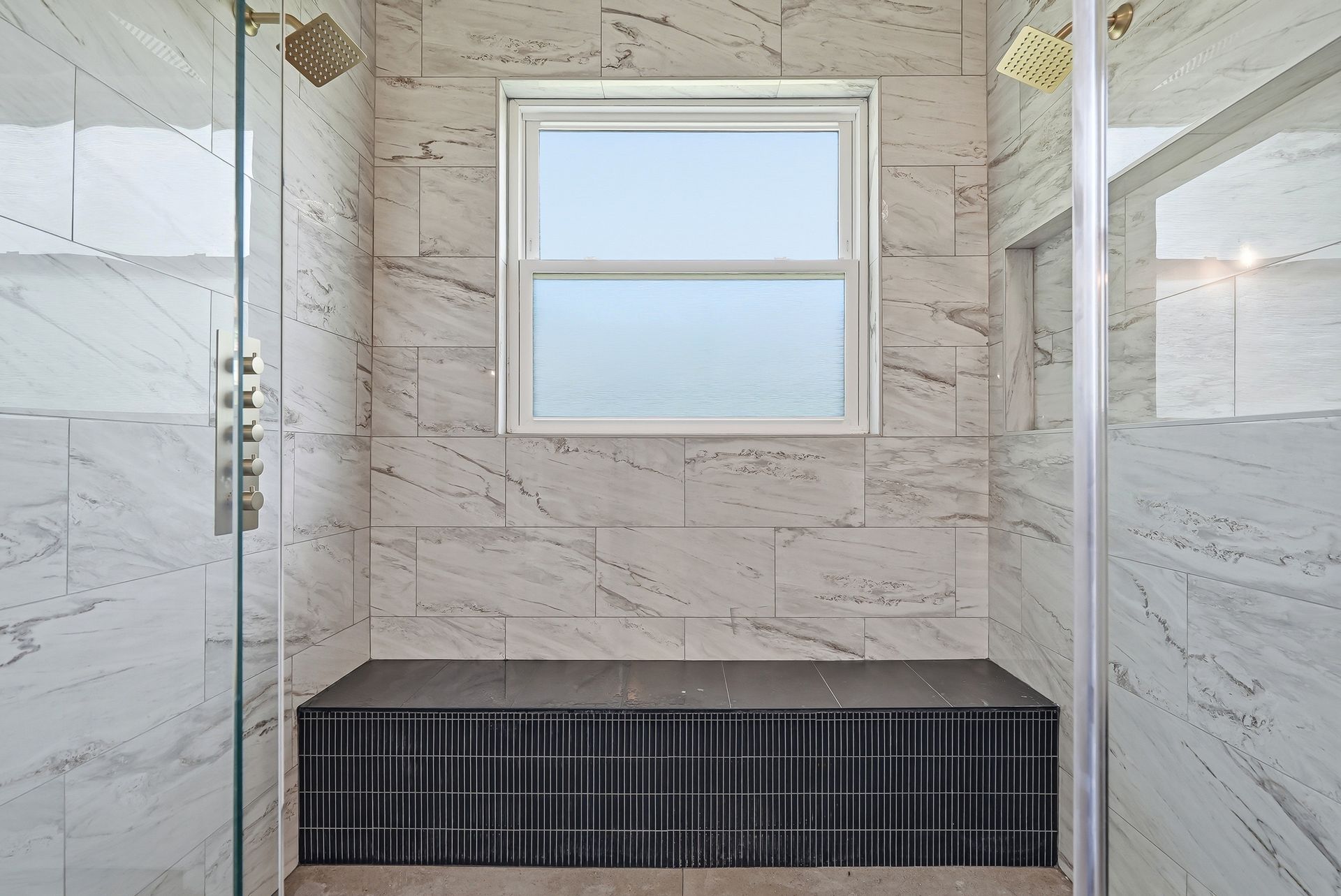 Luxury walk-in shower with dual gold rainfall shower heads, marble-look tile, and built-in tiled bench