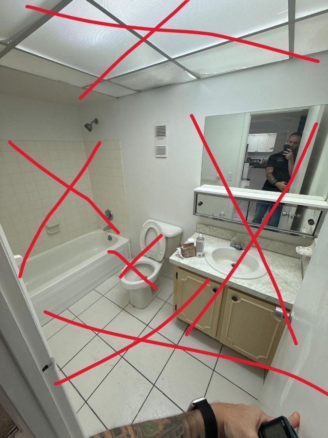 Damaged bathroom with red X marks on walls, ceiling, sink, toilet, and mirror; person visible in mirror.