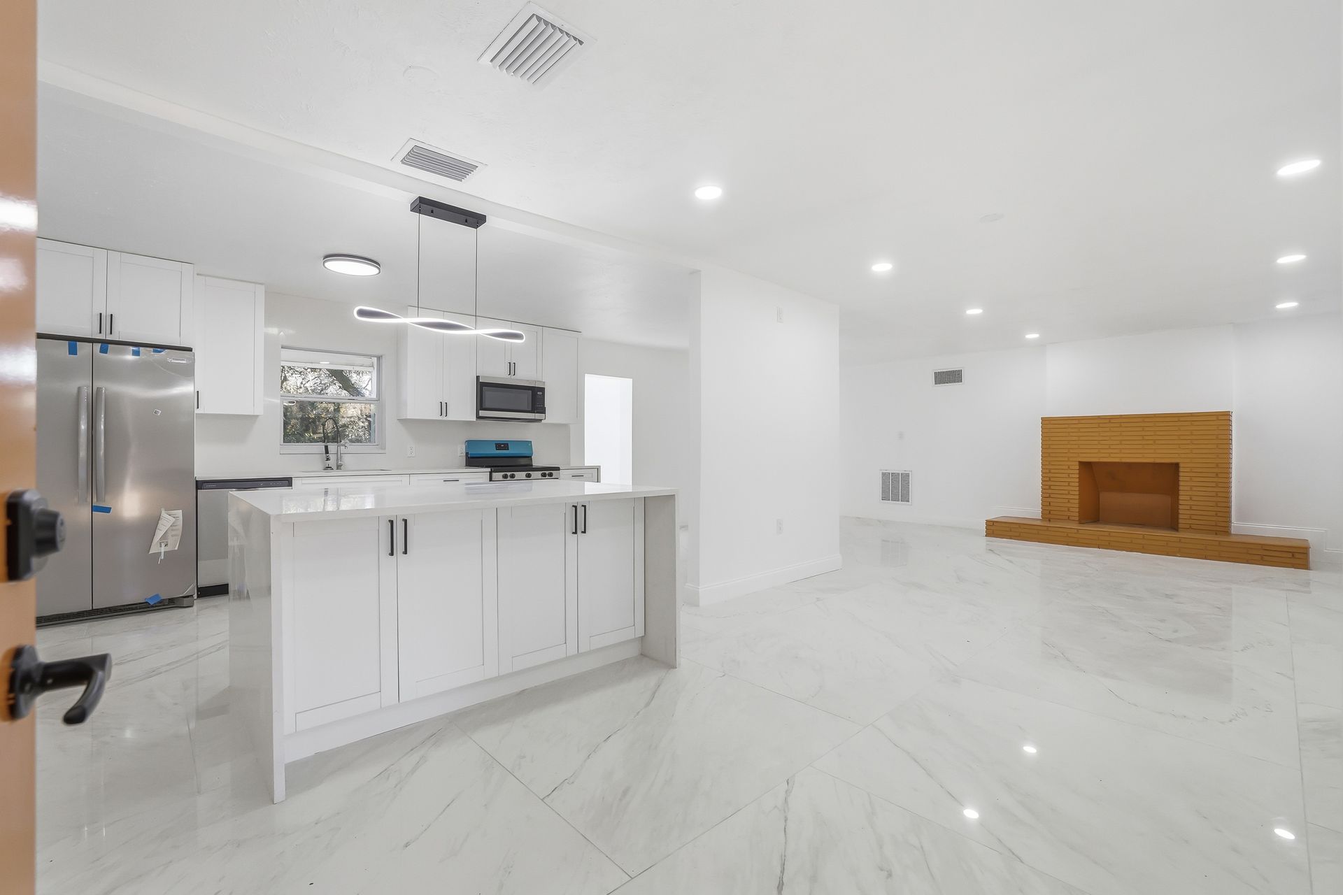 Bright white modern kitchen with island, stainless appliances, and a living room with a fireplace in the background