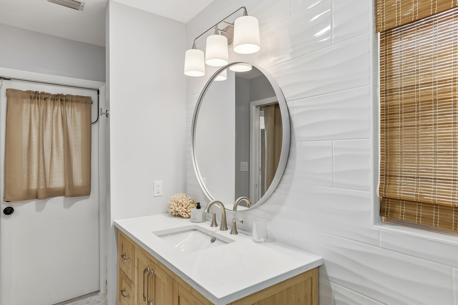 Bright bathroom vanity with round mirror, white tile, wood cabinet, and woven window shade