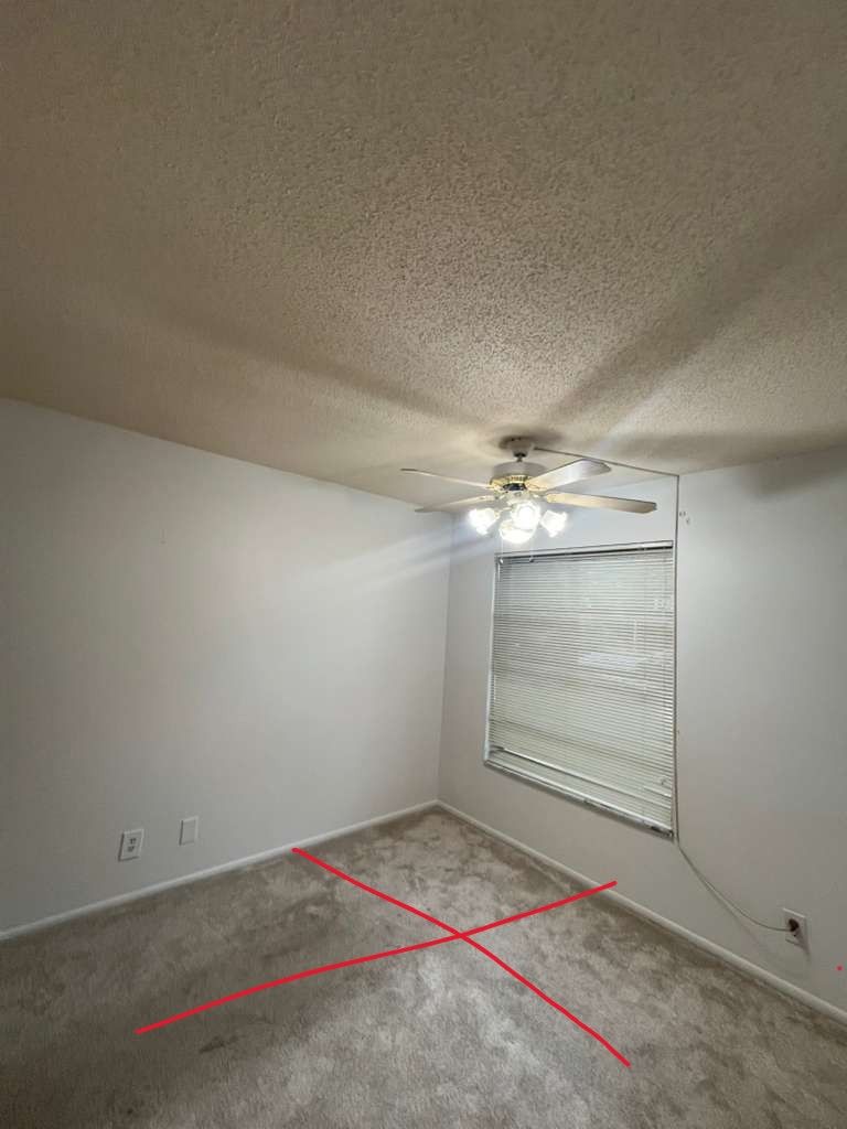 Empty carpeted bedroom with white walls, ceiling fan, and window blinds; red X marked on the floor.