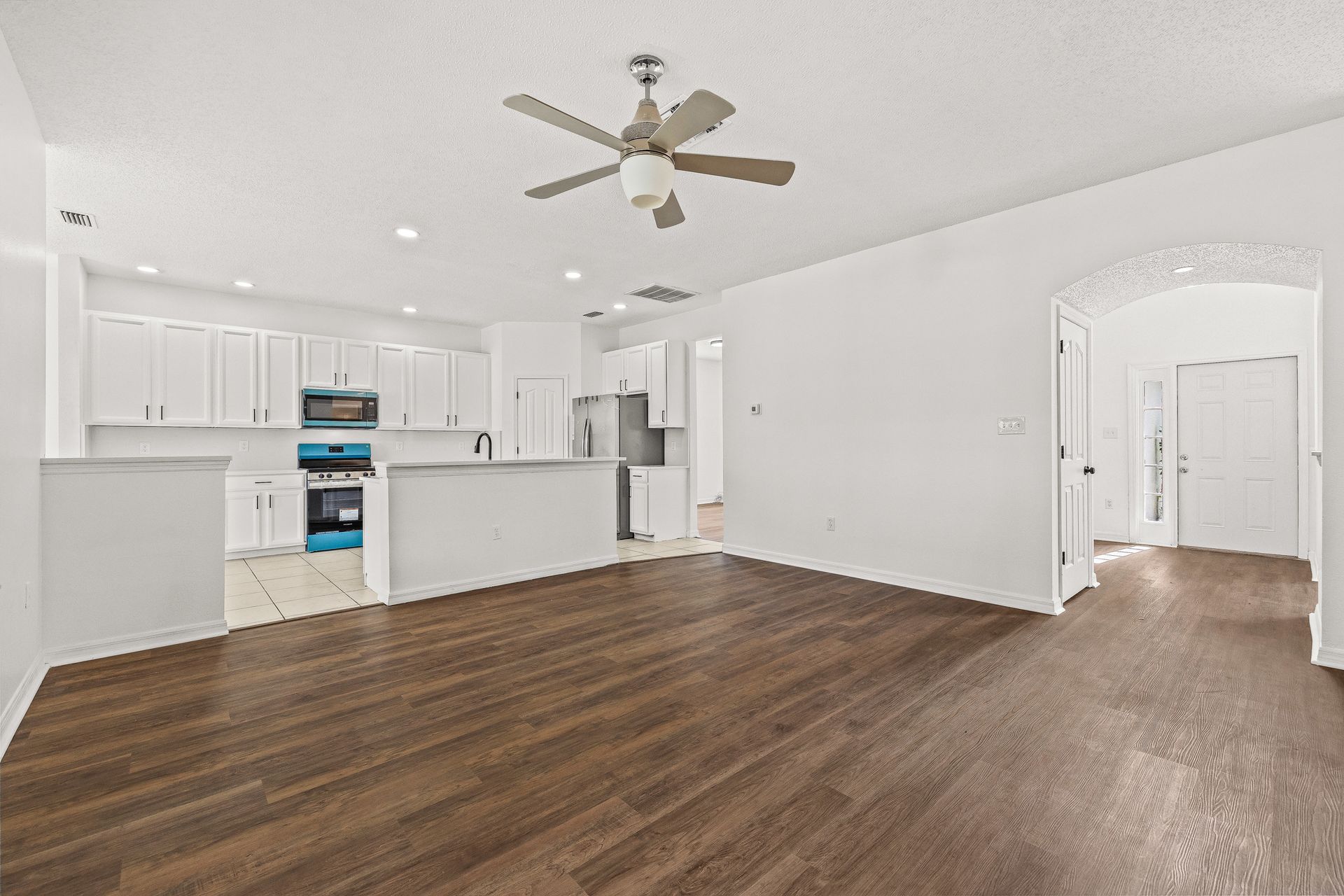 Bright empty open-concept living room and kitchen with white walls and wood floors, ceiling fan, and arched doorway.