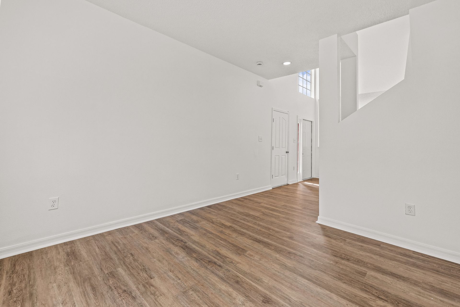 Empty white room with wood floors, high ceiling, and a bright entry hallway
