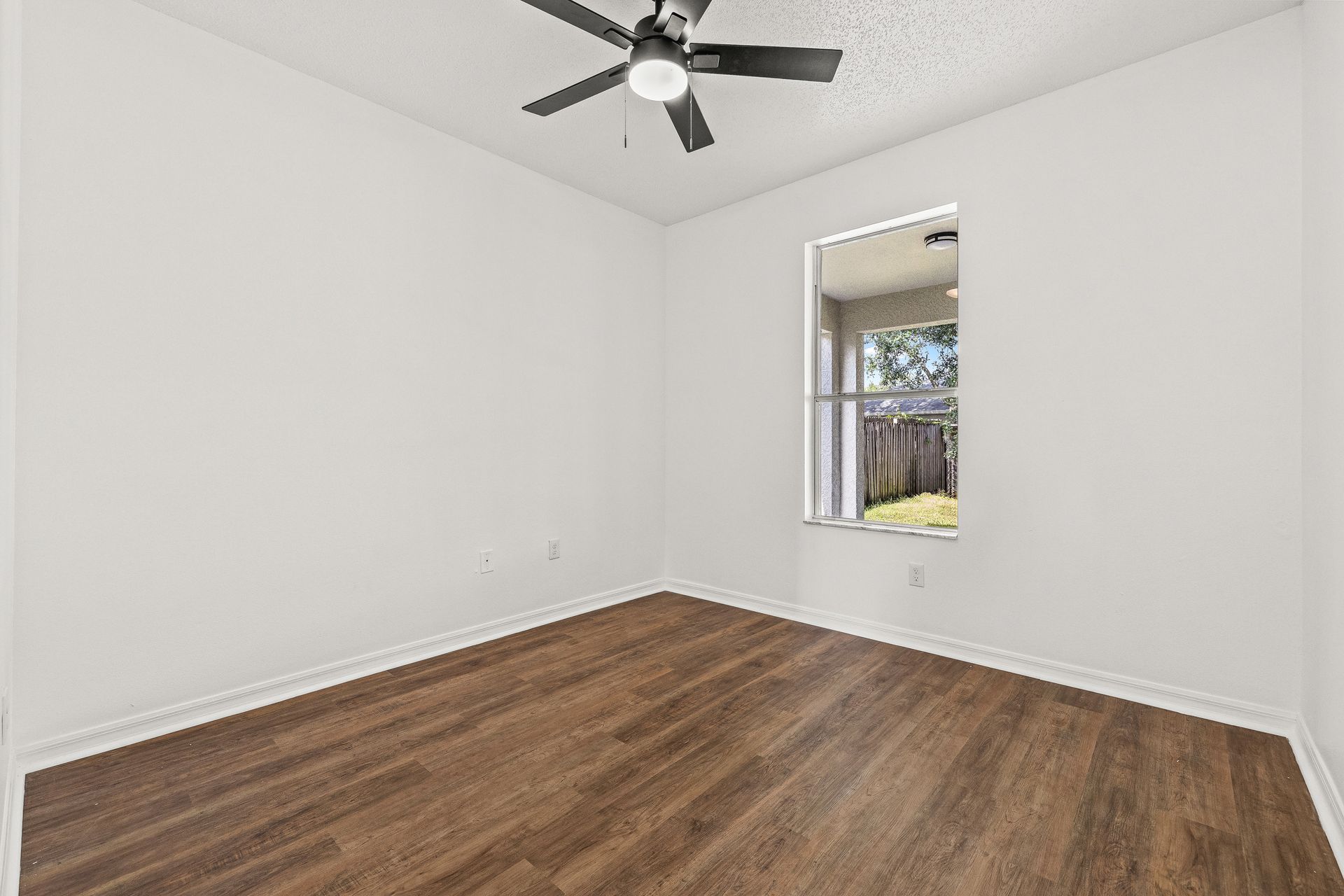 Empty white room with wood floors, ceiling fan, and a small window looking outside