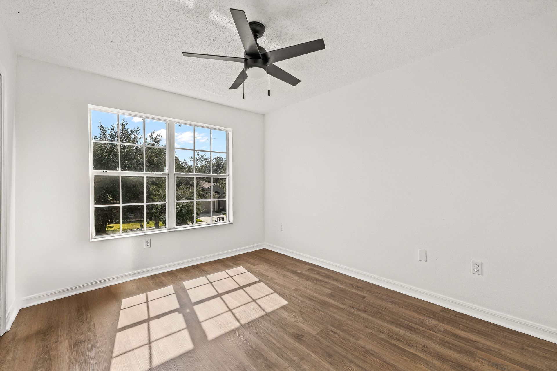 Empty white bedroom with wood floor, ceiling fan, and large window casting sunlight on the floor.
