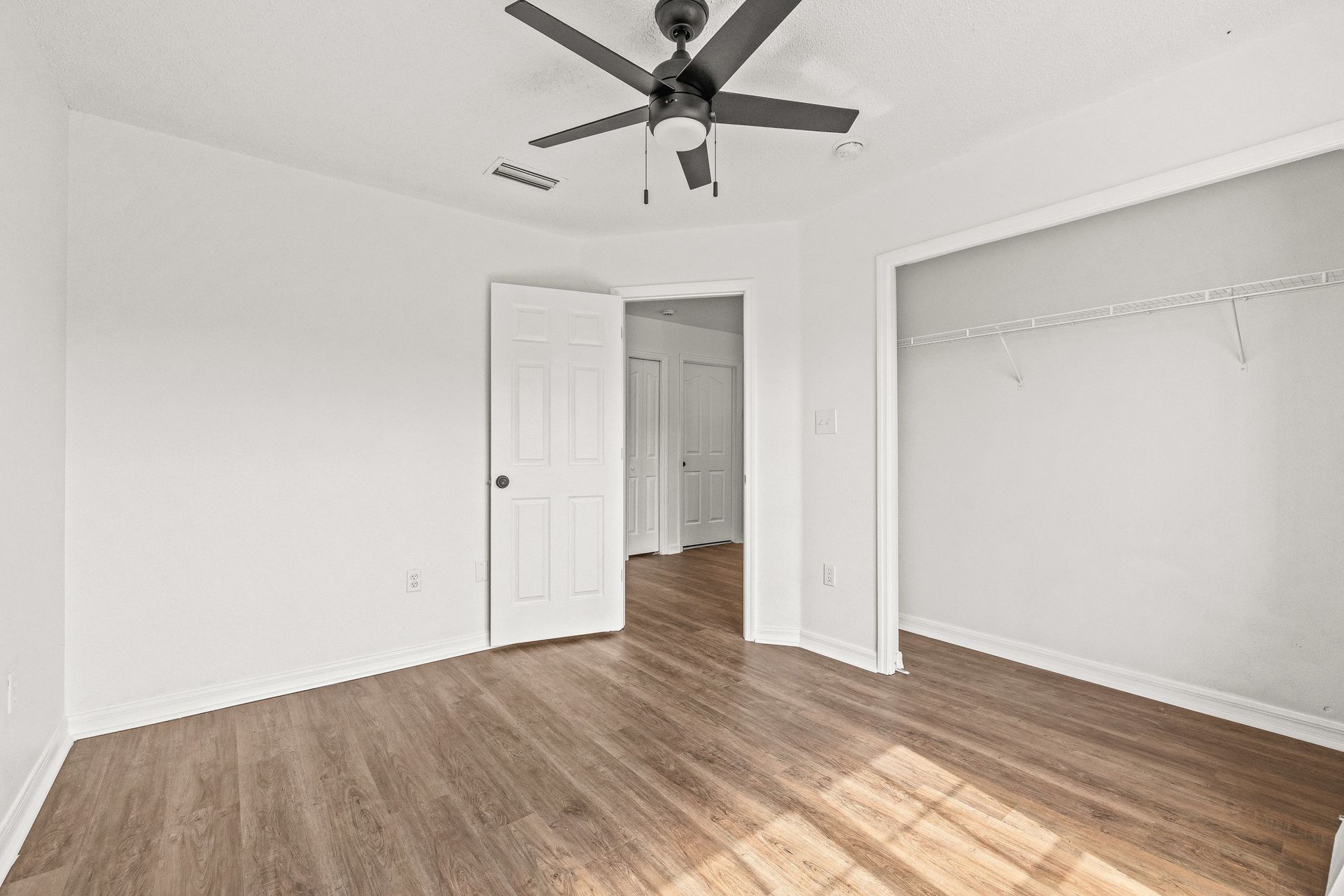 Empty white bedroom with wood flooring, ceiling fan, and open closet doors