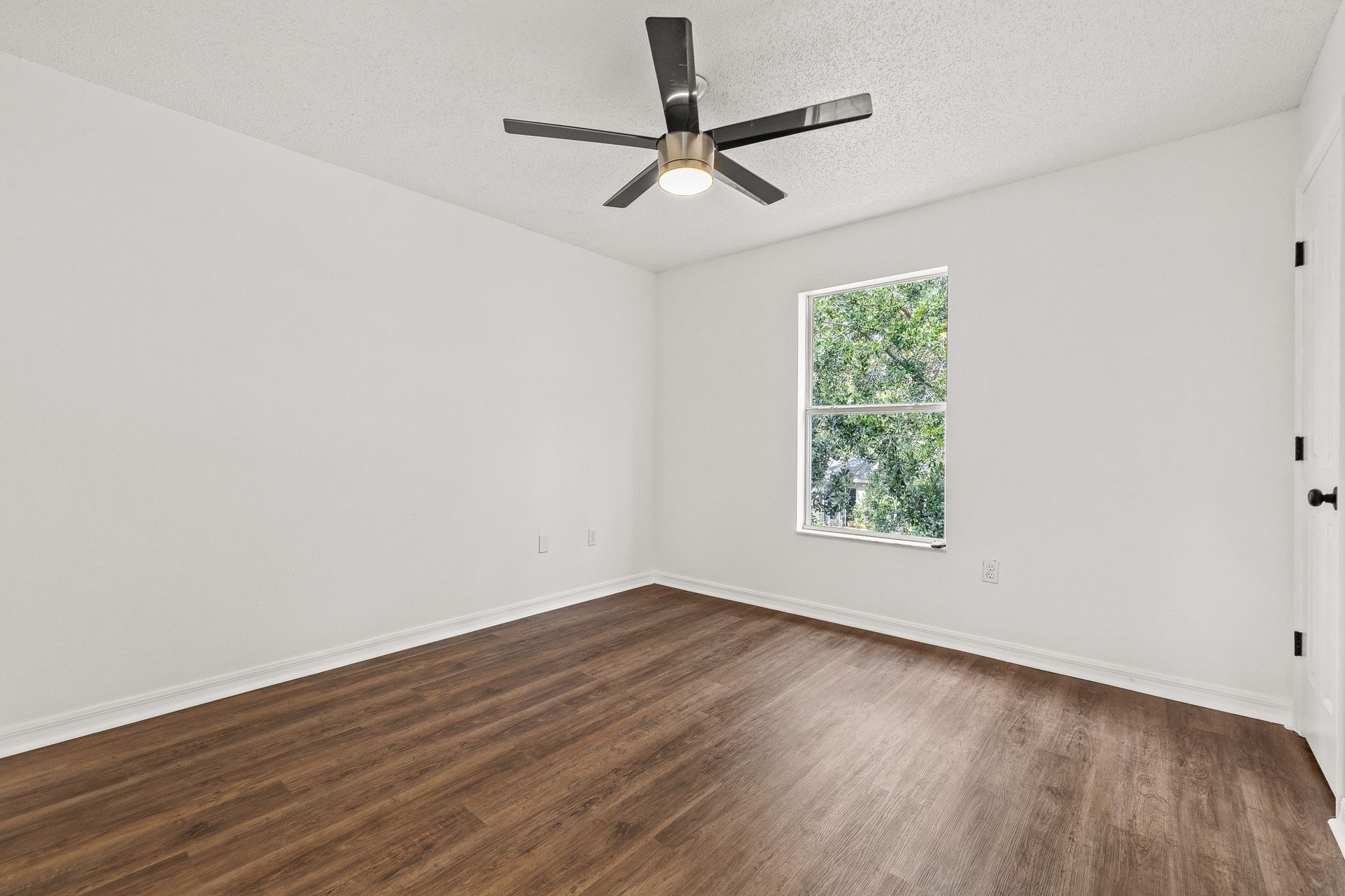 Empty white bedroom with wood flooring, ceiling fan, and a window with greenery outside