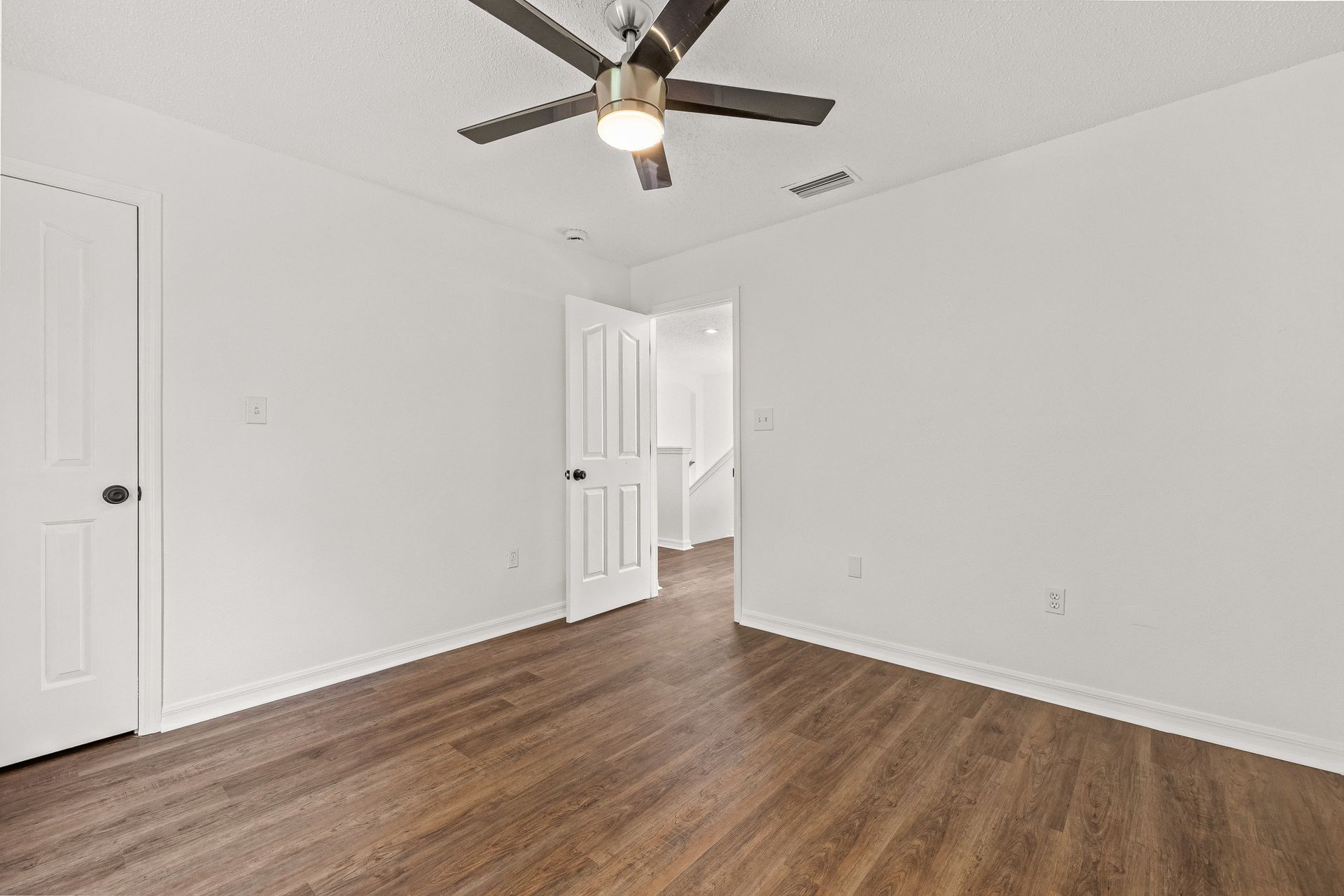 Empty white bedroom with wood flooring, ceiling fan, and open door to a hallway