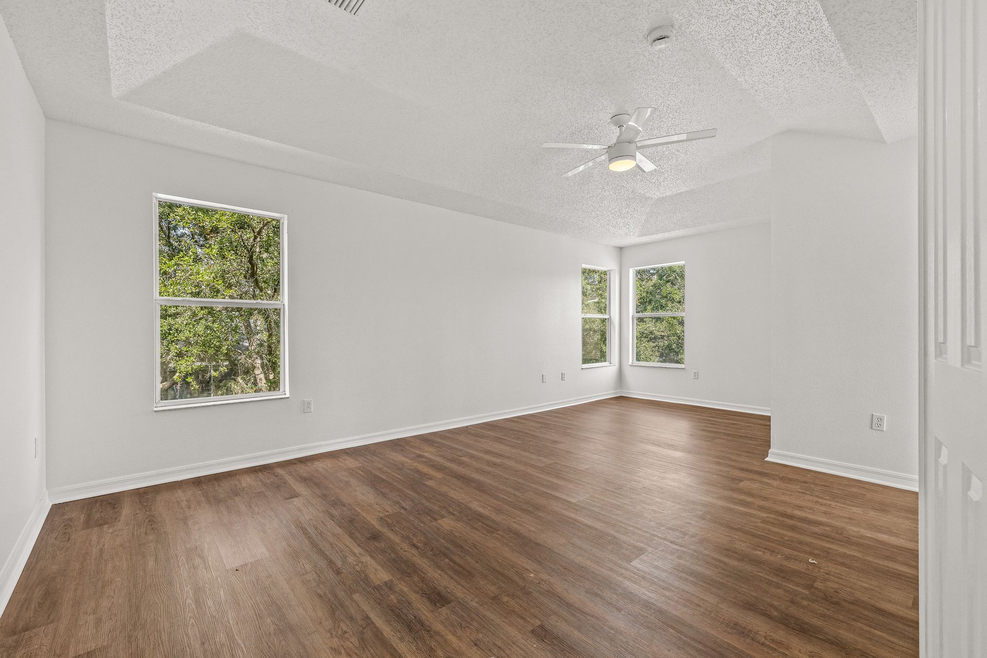 Empty white living room with hardwood floors, a ceiling fan, and windows letting in green outdoor light