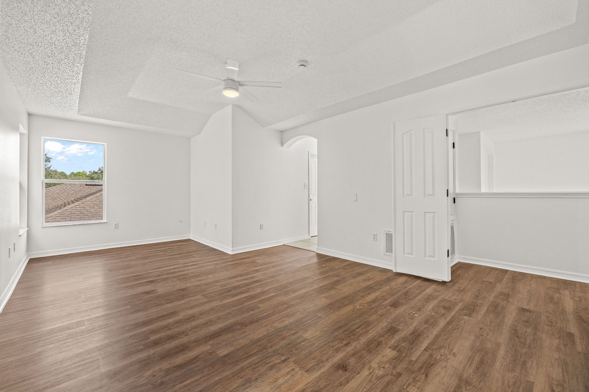 Empty white bedroom with wood floors, ceiling fan, closet doors, and a window overlooking a deck.