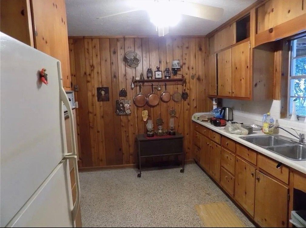 Small rustic kitchen with wood-paneled walls, white fridge, wooden cabinets, sink, and window.