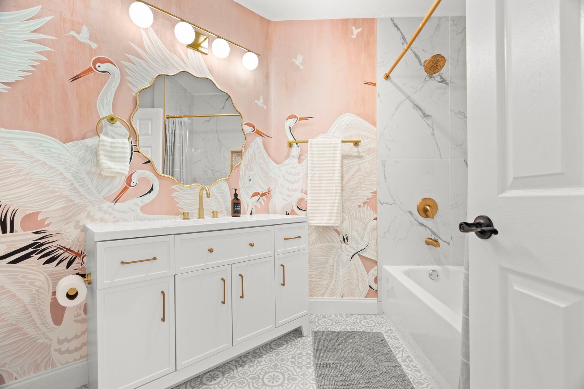Designer bathroom renovation featuring pink crane-pattern wallpaper, a white double-sink vanity, scalloped gold-framed mirror, brass fixtures, patterned floor tile, and a marble-look bathtub surround.