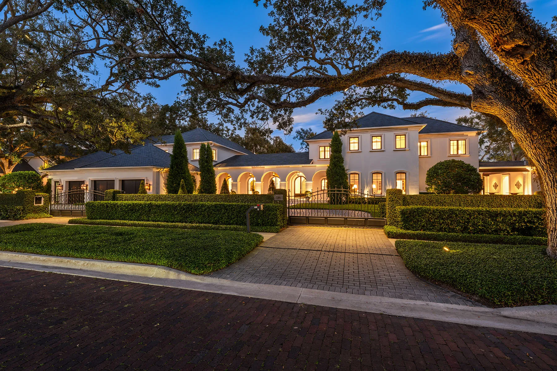 Illuminated luxury mansion at dusk with manicured hedges, large trees, and a lit driveway entrance
