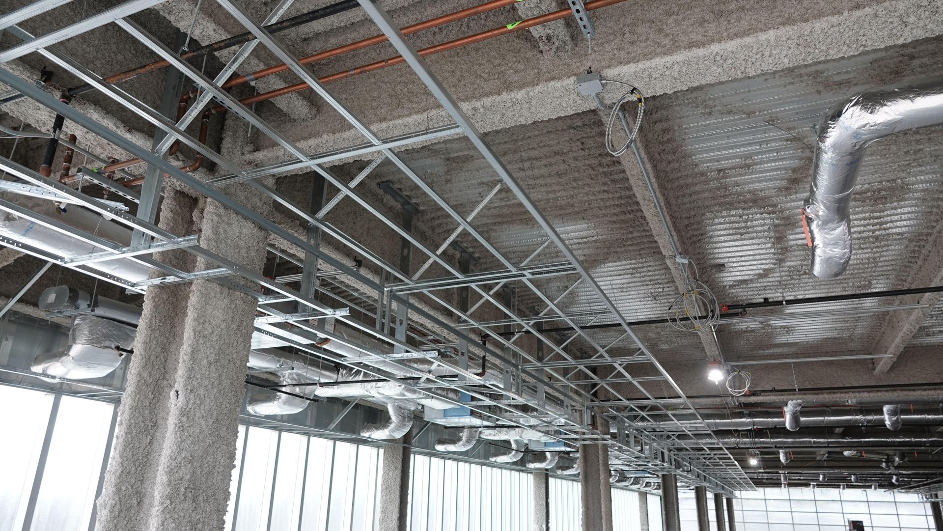 Unfinished industrial ceiling with exposed ducts, pipes, and grid framing above large windows