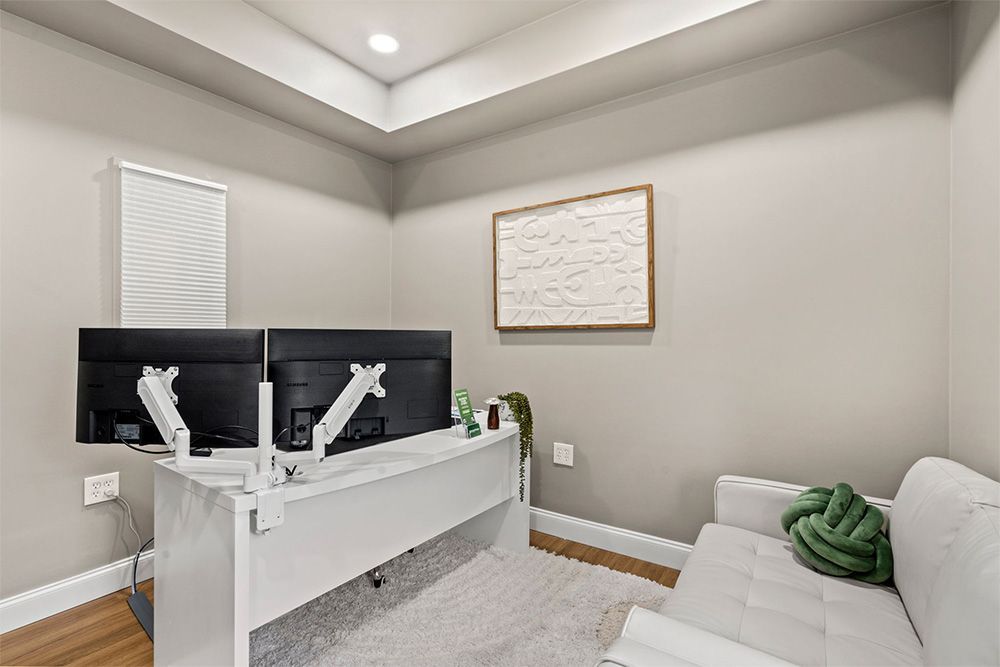 Modern office with dual monitors, white desk, gray walls, framed art, and a white couch with green pillows.