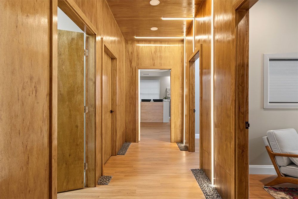 Wood-paneled hallway with recessed lights, open doors, and a chair beside a white wall panel.