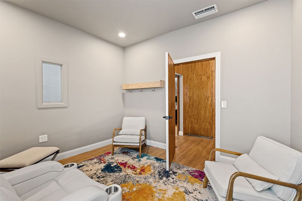 Small white room with chairs, colorful rug, and open door with wooden hallway beyond