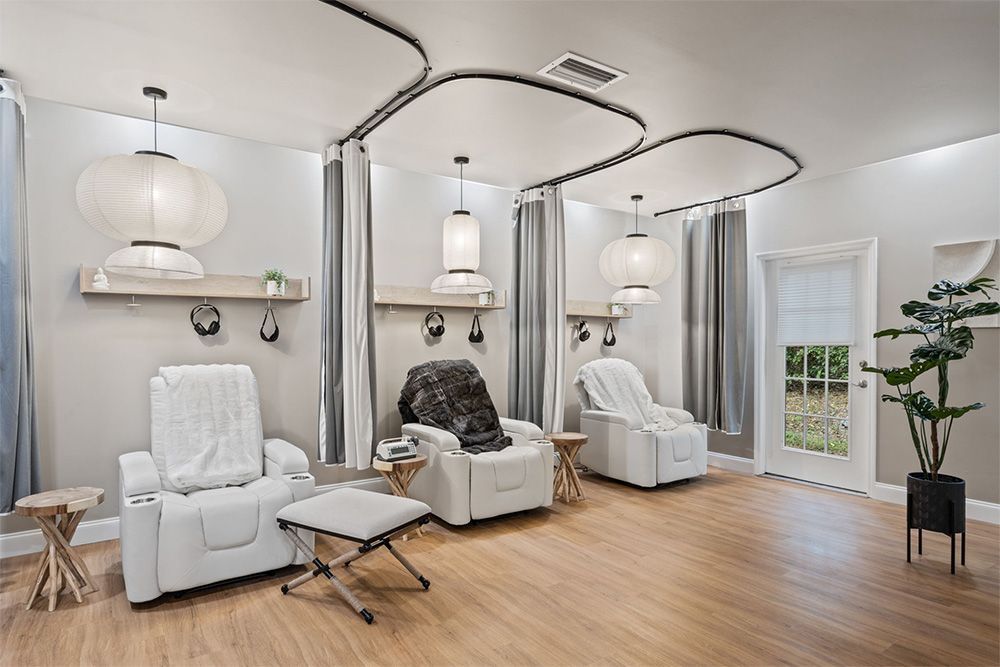 Modern spa treatment room with white recliners, warm wood floors, pendant lights, and a potted plant by the window