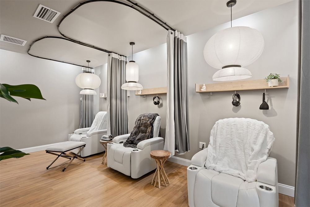 Bright spa treatment room with white recliners, curtains, hanging lamps, and light wood floors