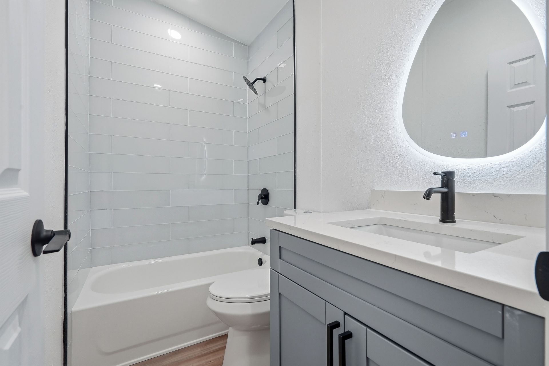 Modern bathroom renovation with white subway tile tub surround, gray shaker-style vanity, quartz countertop, illuminated round mirror, and matte black fixtures by Step Up Home Improvements.
