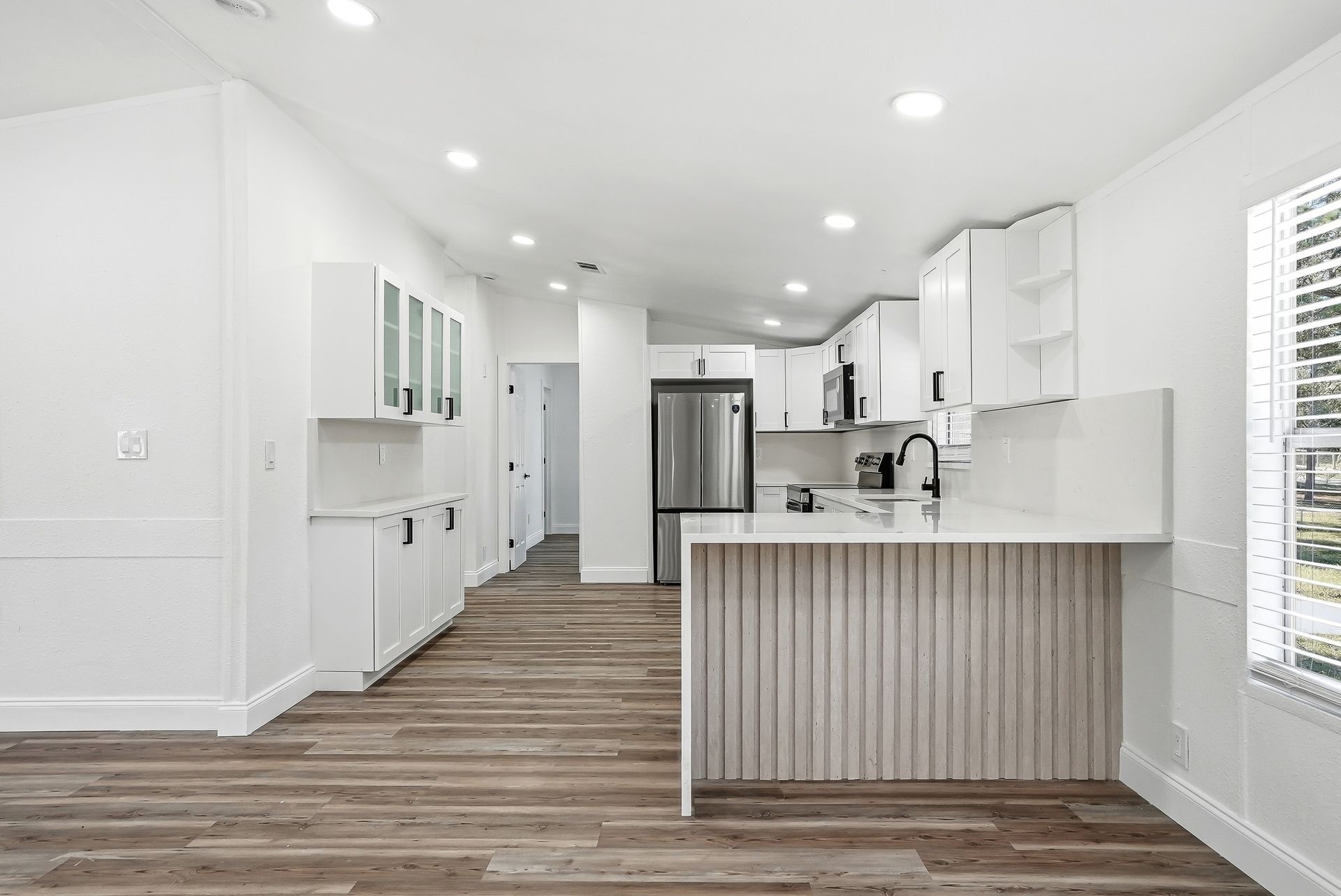Modern white kitchen remodel by Step Up Home Improvements featuring shaker cabinets, quartz countertops, a waterfall-edge peninsula, wood-look flooring, black hardware, and recessed lighting.