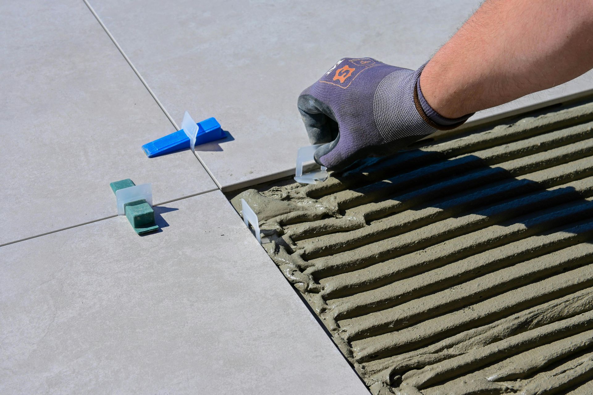 Gloved hand spreading adhesive on floor tiles beside blue and green spacers