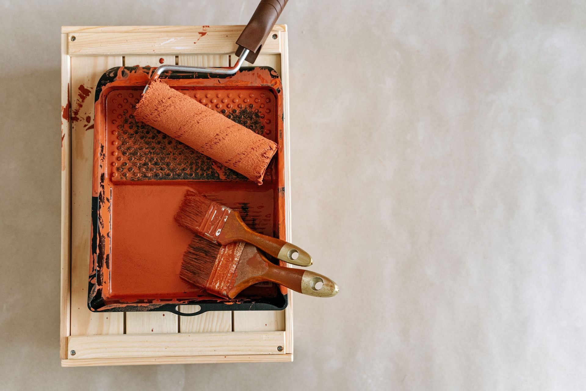 Paint tray with orange paint, roller, and brush on a wooden frame against a light background