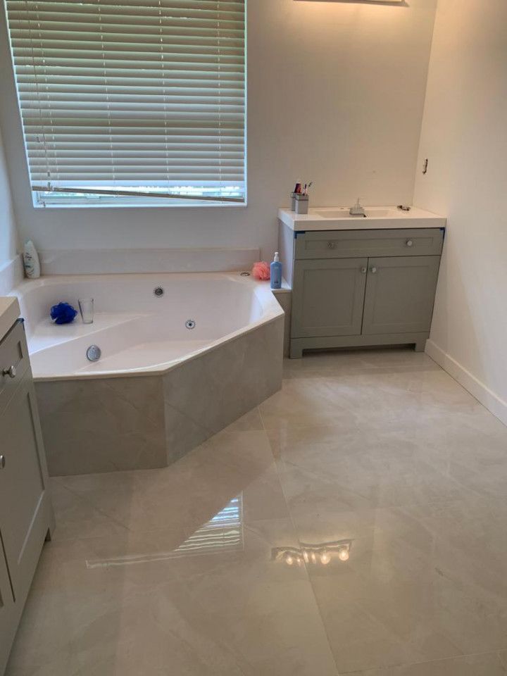 Bright bathroom with corner soaking tub, gray vanity, window blinds, and beige tile floor