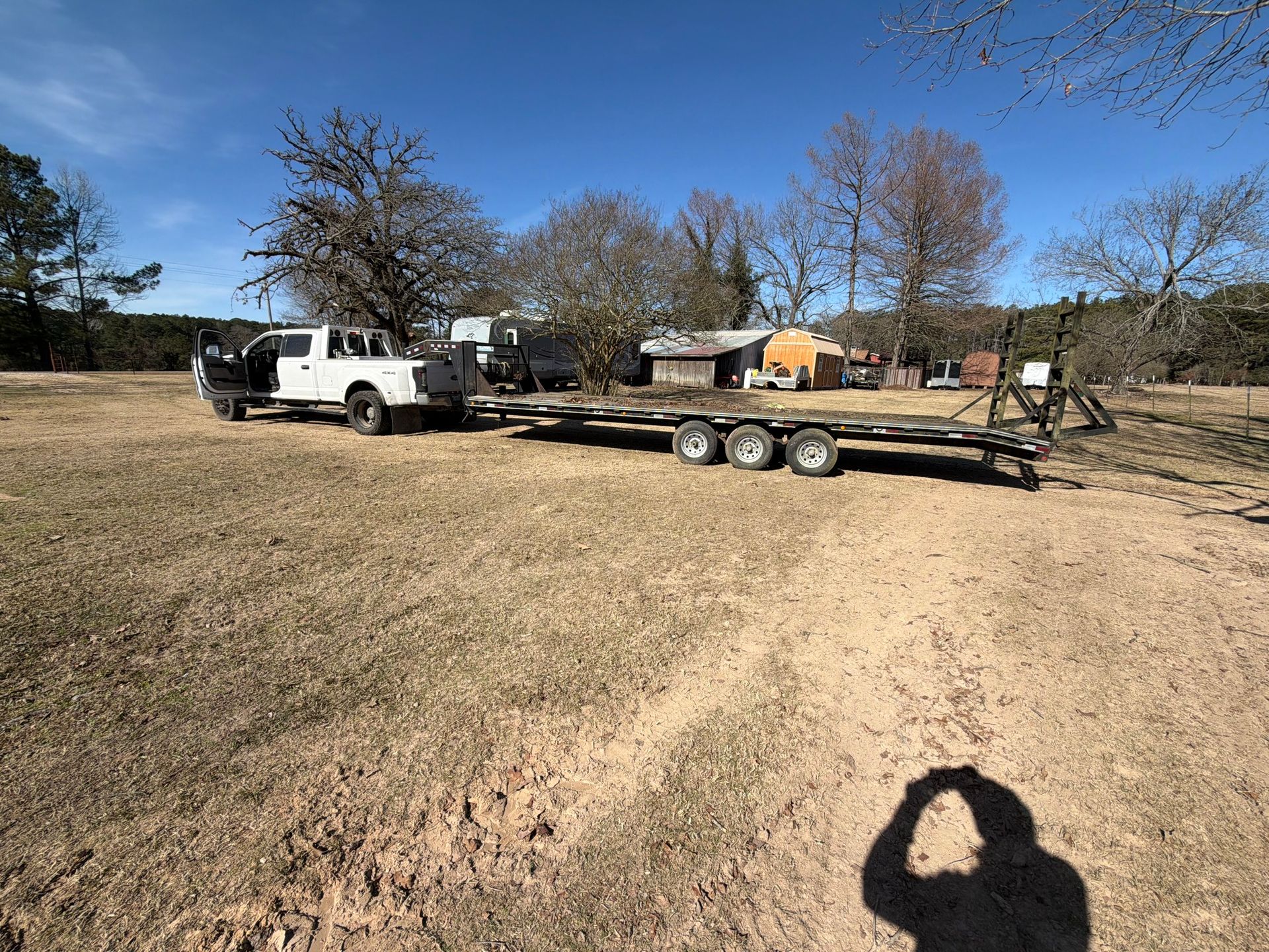 A white pickup truck pulling a long, empty flatbed trailer in a dry, open field on a sunny day.