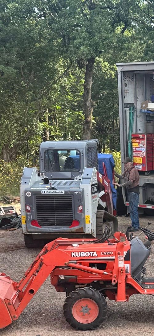 A person works near a grey skid steer and an orange tractor parked in a wooded area next to a trailer.