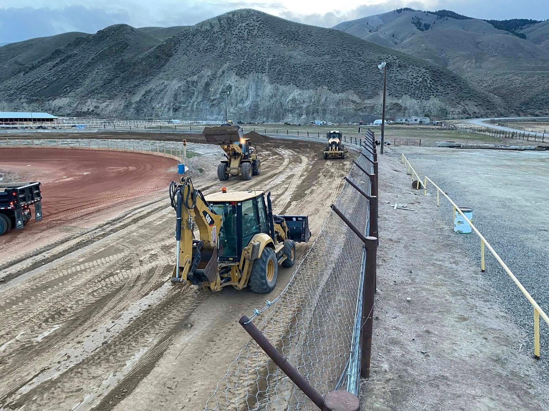 Yellow construction equipment works on a dirt track in front of a mountain range under a cloudy sky.