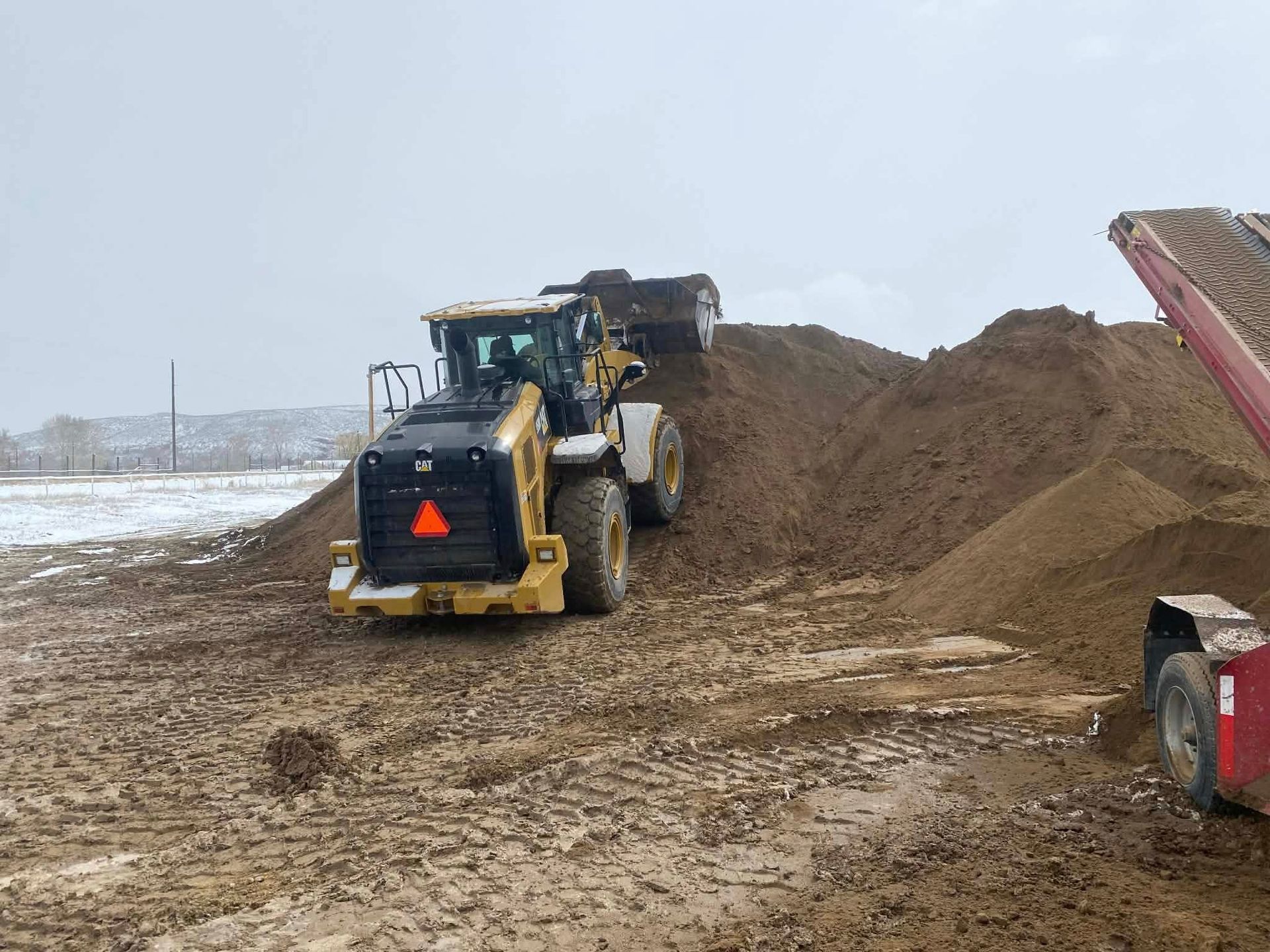 A yellow front loader moves soil near a conveyor system on a muddy, snow-dusted construction site.