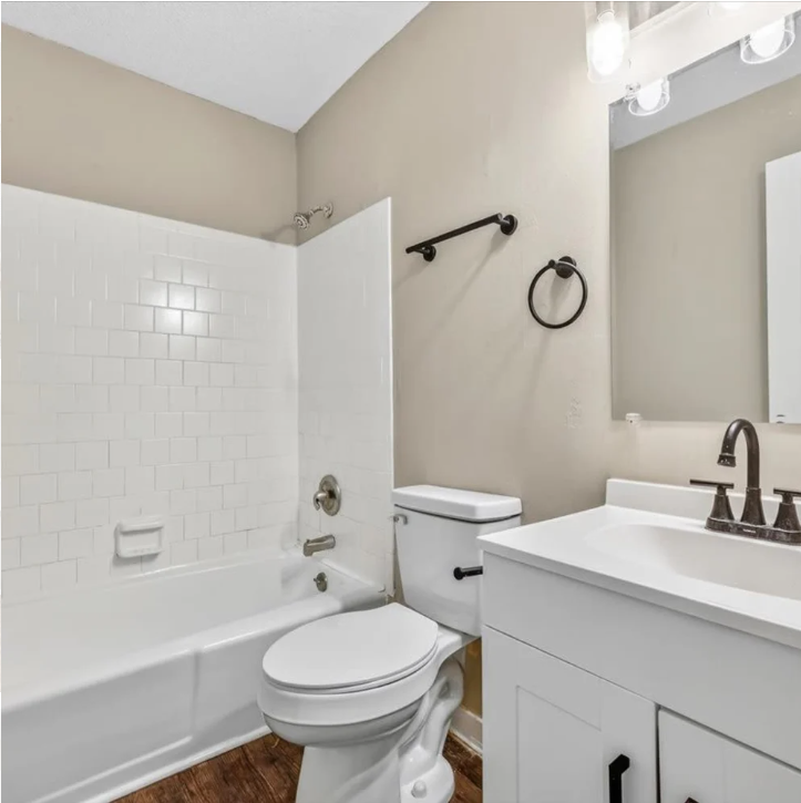 Small beige bathroom with white tub-shower, toilet, vanity, mirror, and towel ring.