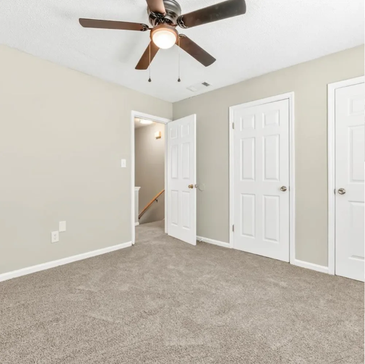 Empty beige bedroom with carpet, ceiling fan, and three white closet doors