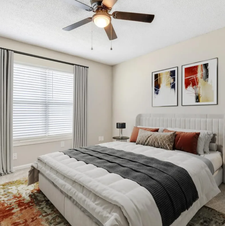 Bright bedroom with a gray bed, abstract wall art, ceiling fan, and large window with curtains