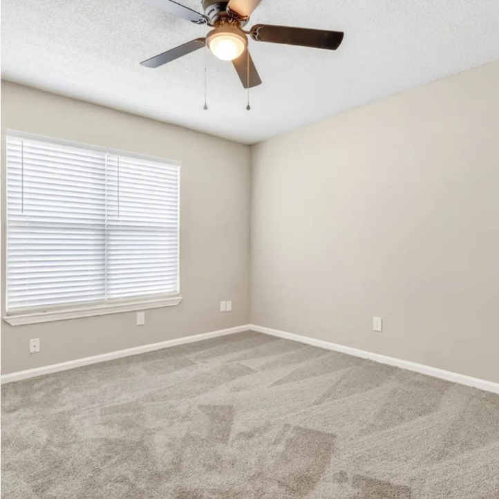 Empty beige bedroom with carpet, window blinds, and a ceiling fan with light.