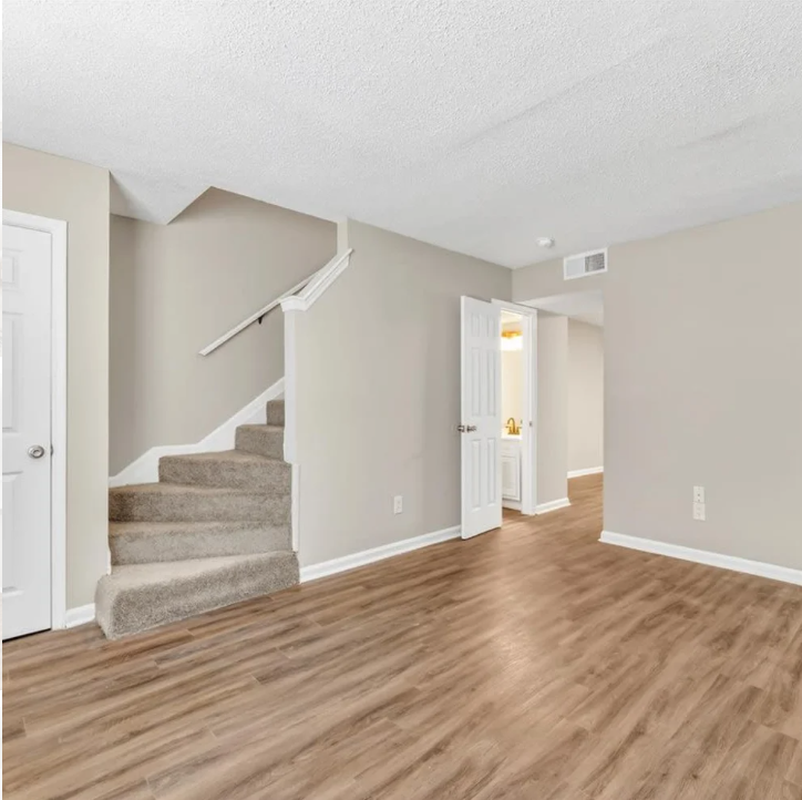 Empty beige living room with wood floors, carpeted stairs, and an open doorway to a bright hallway