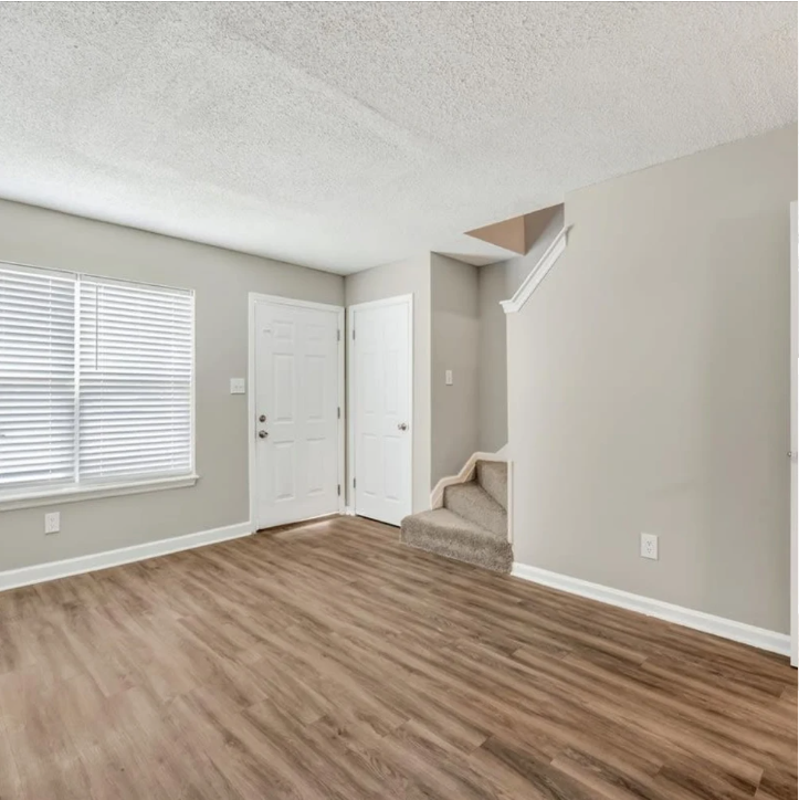 Empty living room with gray walls, wood flooring, white door, and staircase alcove