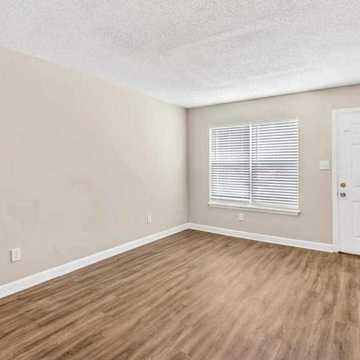 Empty beige living room with wood-look flooring, white trim, and a window with blinds.