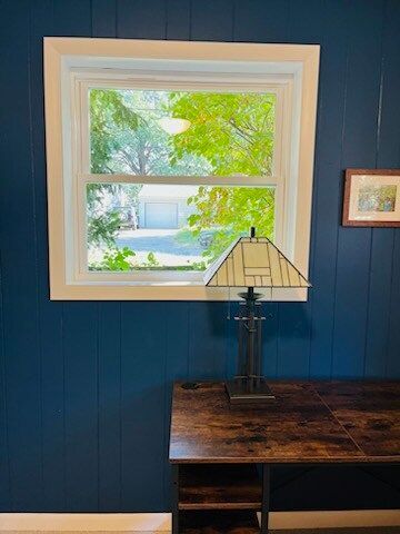 Blue paneled wall with a window, desk, and lamp. White window trim. Bright green outside.