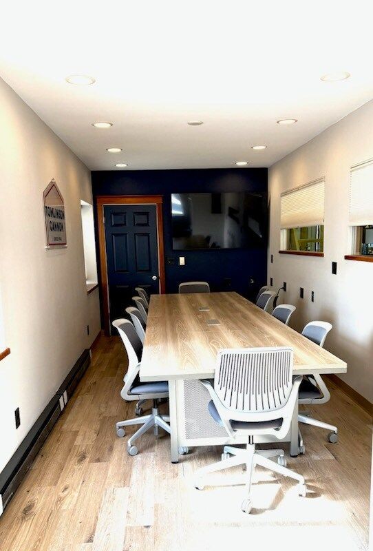 Conference room with a large wooden table, gray chairs, and a TV on a dark blue wall.