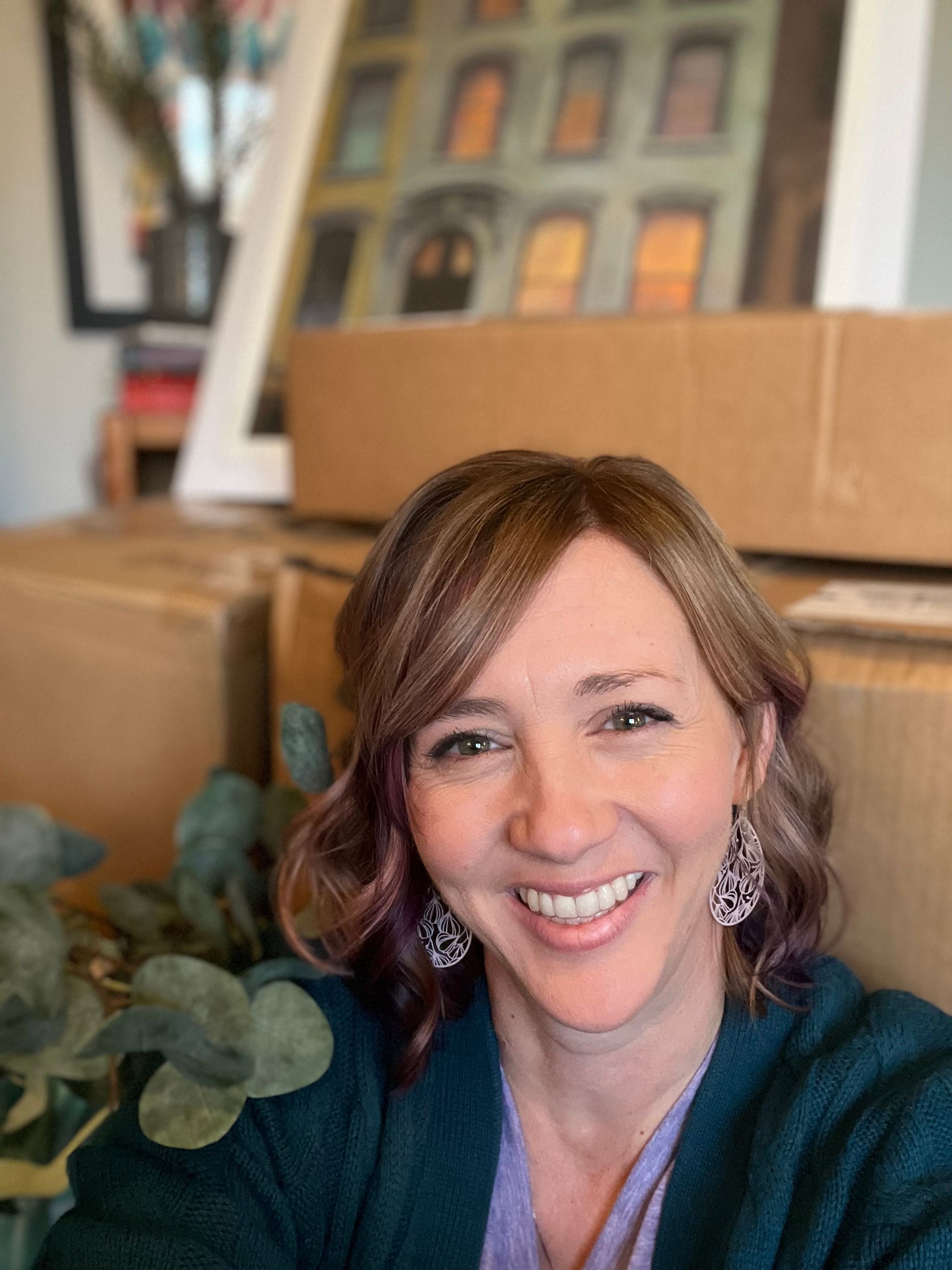 Woman with brown hair and silver earrings smiles, boxes and artwork in background.
