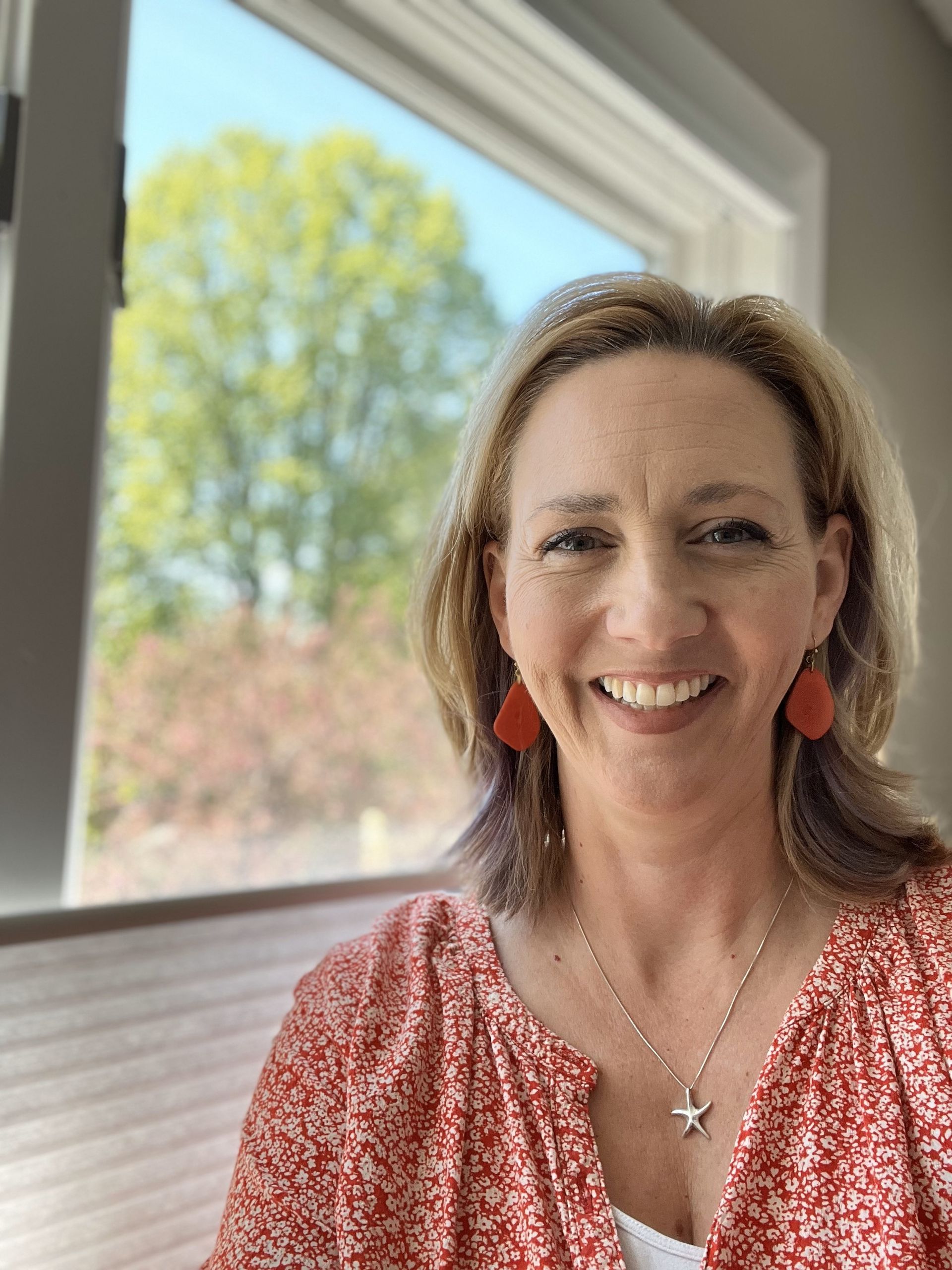 Woman smiling, wearing red blouse and earrings, in front of a window overlooking a tree with green leaves.