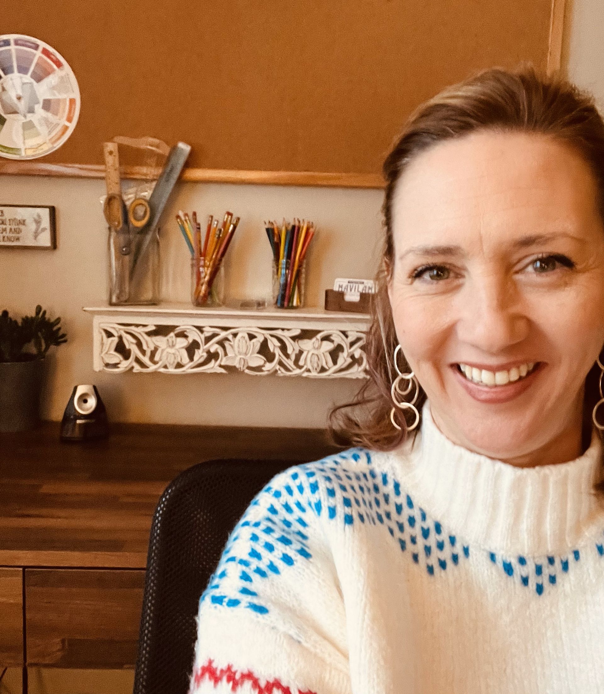 Woman smiling, wearing white sweater, desk with art supplies in background.