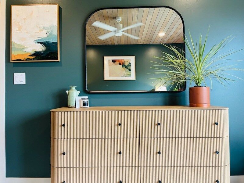 Teal wall with wooden dresser, arched mirror, art, and potted plant.