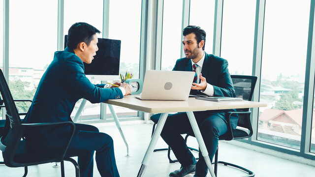 Two men are sitting at a table with a laptop and talking to each other.