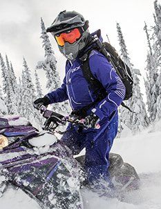 Personne sur une motoneige dans des montagnes enneigées, portant une combinaison et un casque violets.