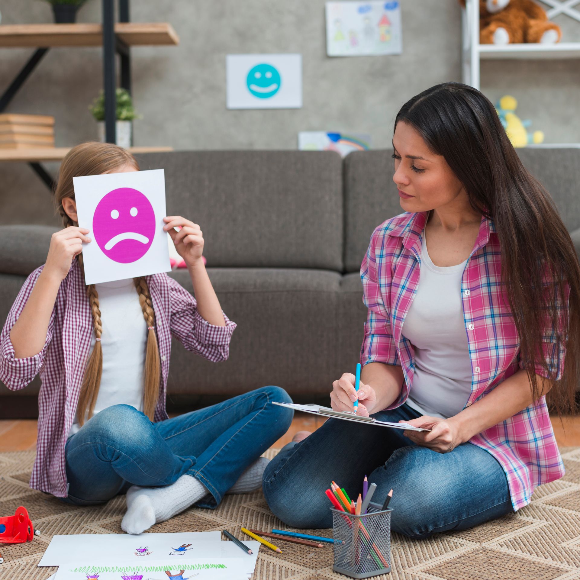 A woman is sitting on the floor with a child holding a sad face in front of her face.