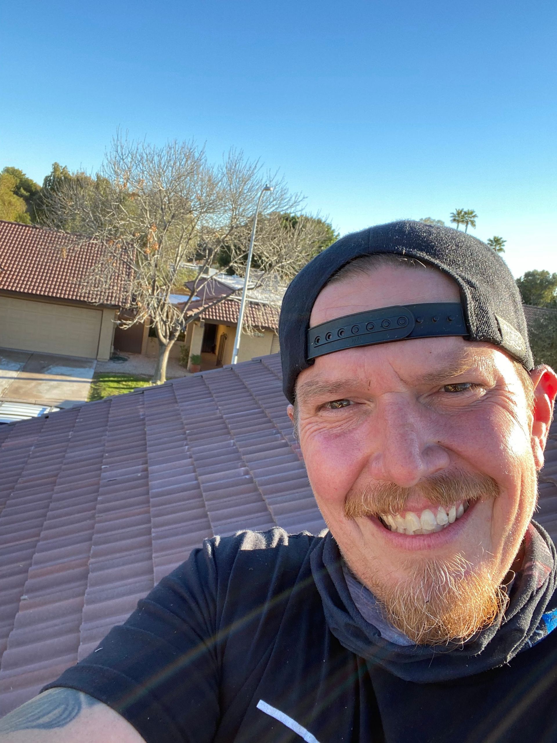 Man on rooftop smiles, wearing a cap, with buildings and trees in background.